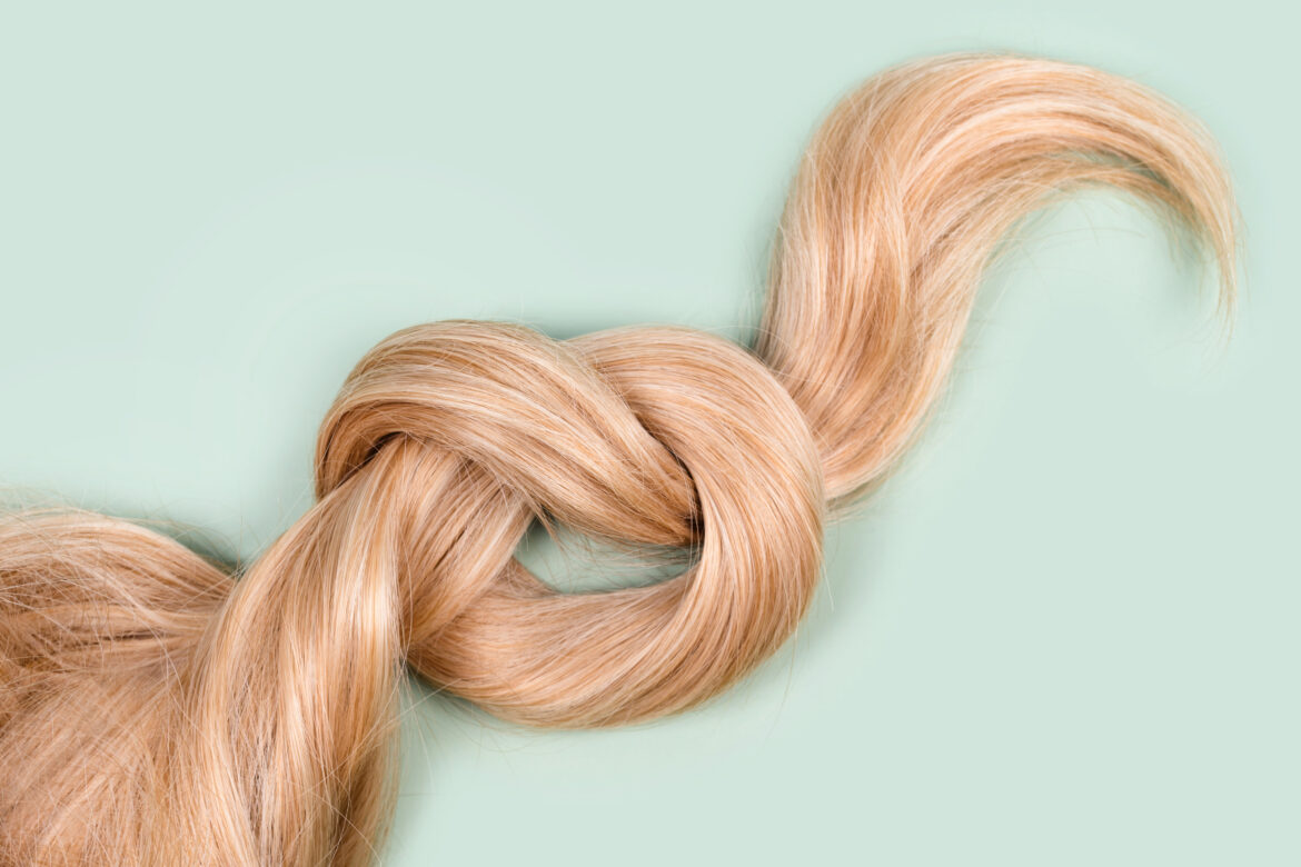 Blonde hair lock tied in knot. Strand of honey blonde hair on mint background, top view. Hairdresser service, hair strength, haircut, hairstyle, dying or coloring, hair extension, treatment concept. Farbowanie włosów na blond – wszystko, co powinnaś wiedzieć, zanim zdecydujesz się na koloryzację